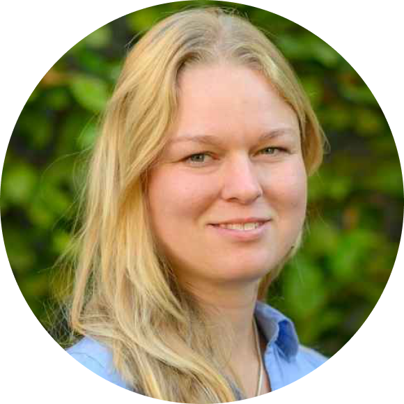 Eine Frau mit langen blonden Haaren, die ein hellblaues Hemd trägt, lächelt in die Kamera. Der Hintergrund besteht aus unscharfem Grün.