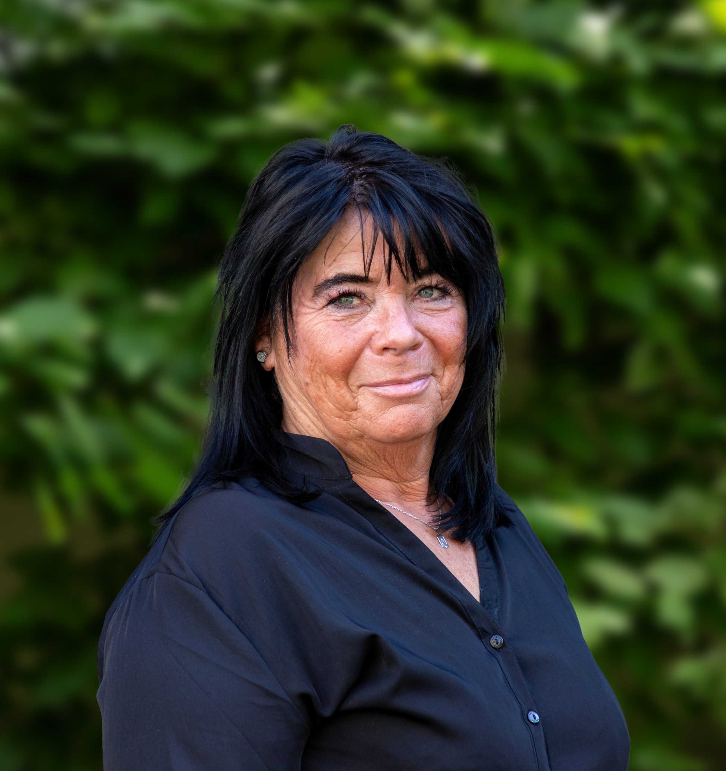 Eine Frau mit glattem schwarzem Haar und schwarzem Hemd steht im Freien vor grünem Laub und lächelt leicht in die Kamera.