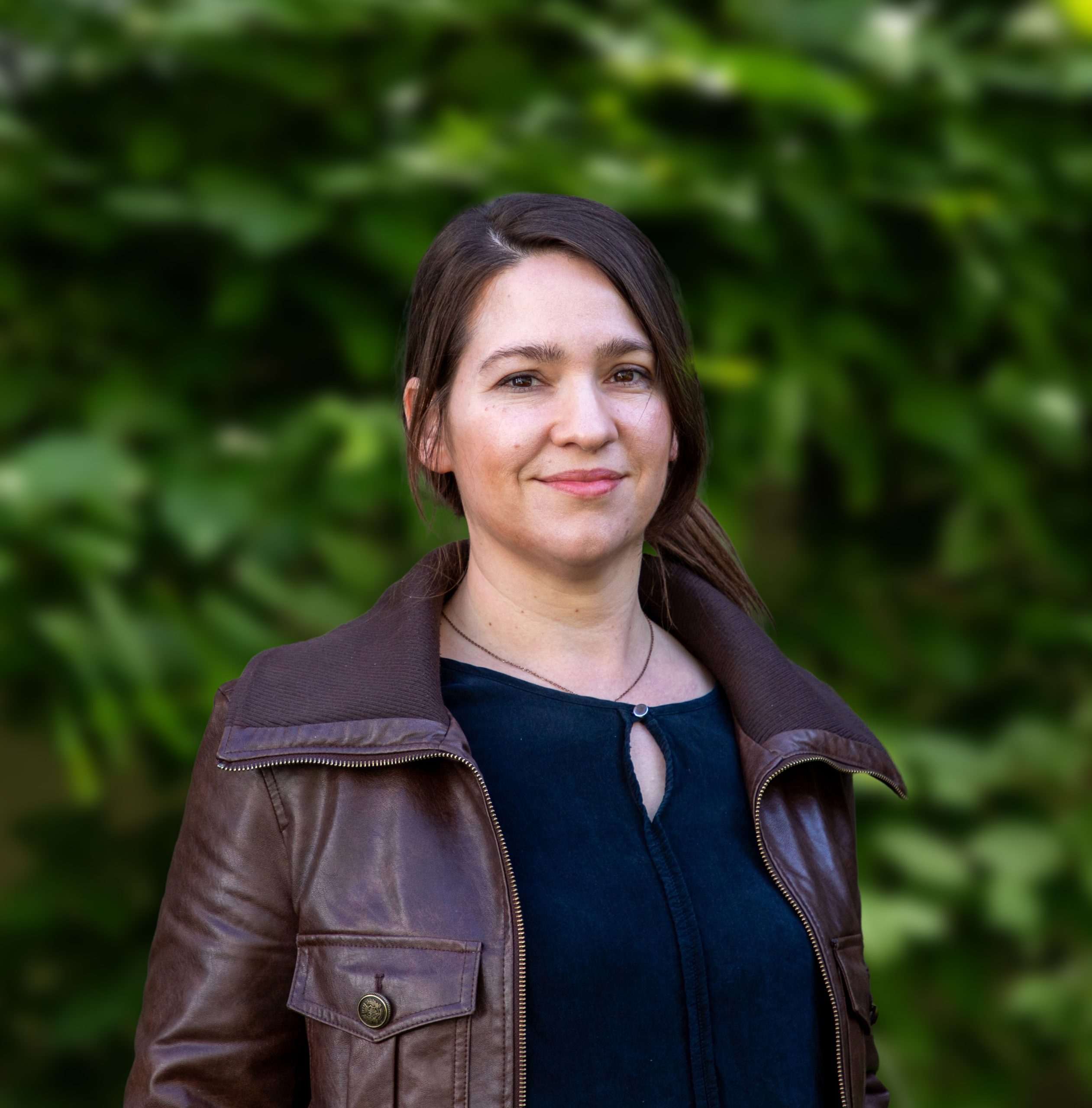 Eine Frau mit langen braunen Haaren, die eine braune Lederjacke und ein dunkles Hemd trägt, steht im Freien vor grünem Blattwerk und lächelt sanft in die Kamera.