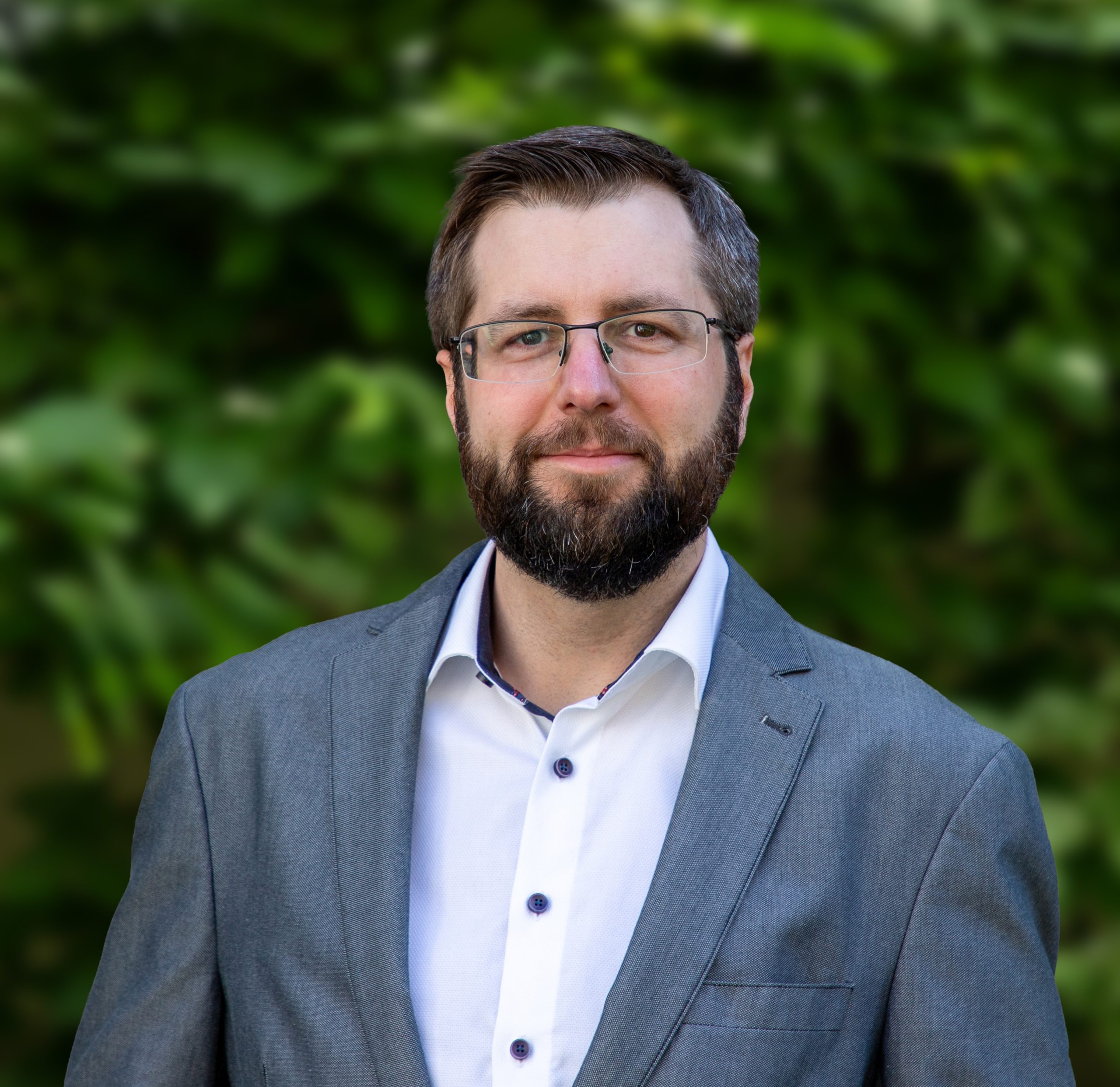 Ein Mann mit kurzen braunen Haaren, Brille und Bart trägt eine graue Anzugsjacke über einem weißen Hemd mit Kragen und steht im Freien vor einem unscharfen grünen Blätterhintergrund.