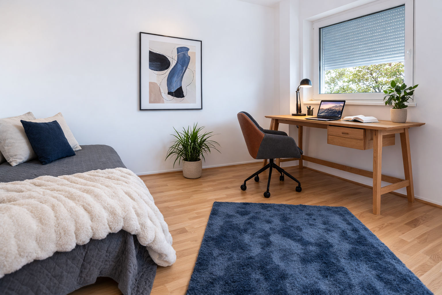 Ein modernes, aufgeräumtes Schlafzimmer mit einem Holzschreibtisch und einem Bürostuhl am Fenster, einem Laptop und einer Lampe auf dem Schreibtisch, einem Bett mit einer grauen Decke und Kissen, einer Pflanze, einem blauen Teppich und abstrakter Wandkunst.