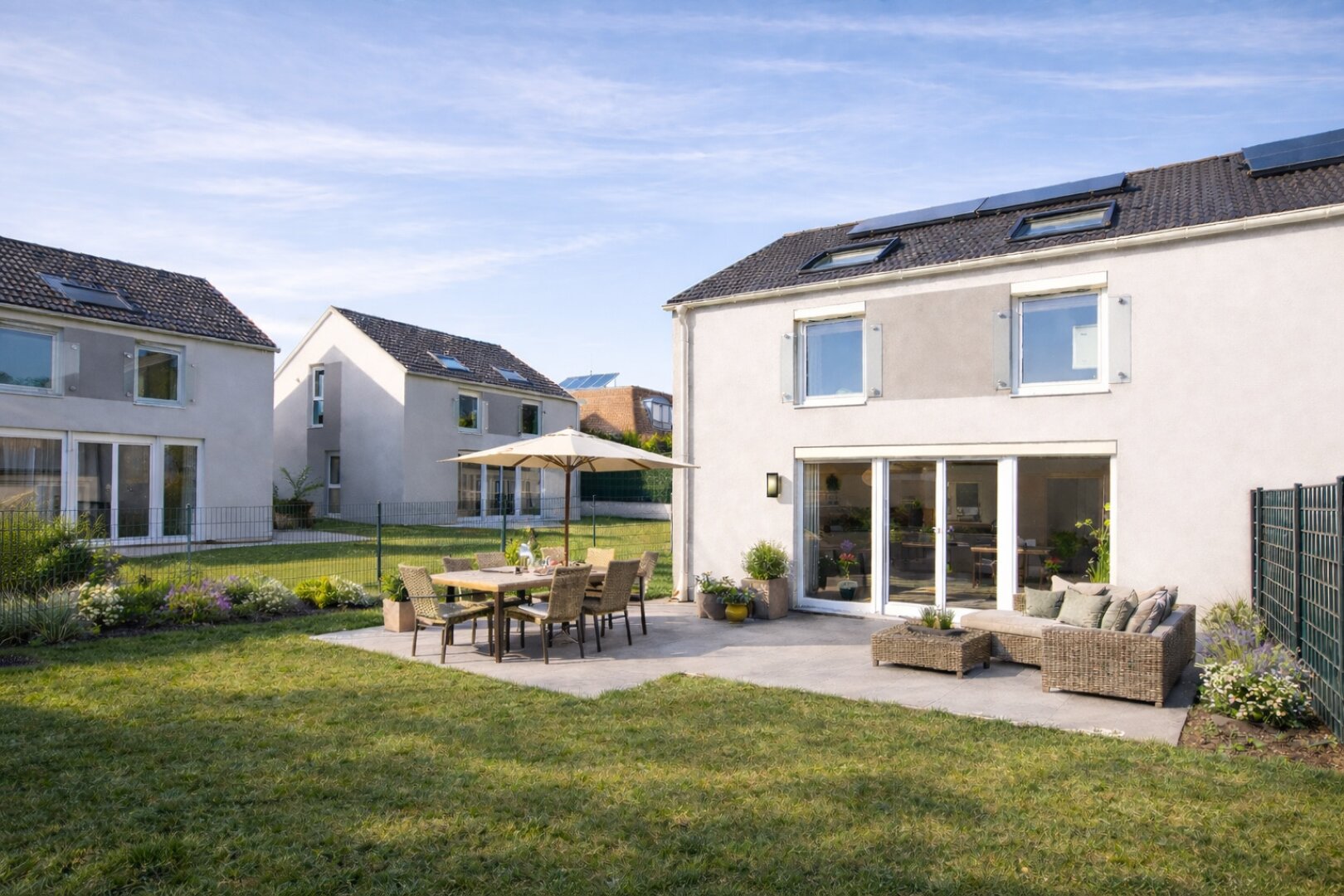 Ein modernes Haus mit großen Fenstern und Sonnenkollektoren steht neben einer Terrasse mit einem Esstisch, Stühlen und einer Couchgarnitur im Freien. Der Hof ist grasbewachsen und mit Blumenbeeten bepflanzt, und im Hintergrund sind ähnliche Häuser zu sehen.