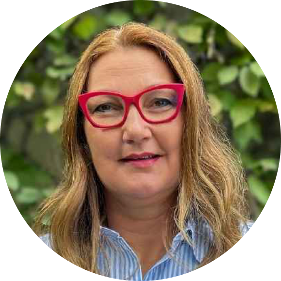 Eine Frau mit langen, hellbraunen Haaren und einer roten Brille lächelt leicht. Sie trägt ein blau-weiß gestreiftes Hemd, im Hintergrund ist grünes Laub zu sehen.