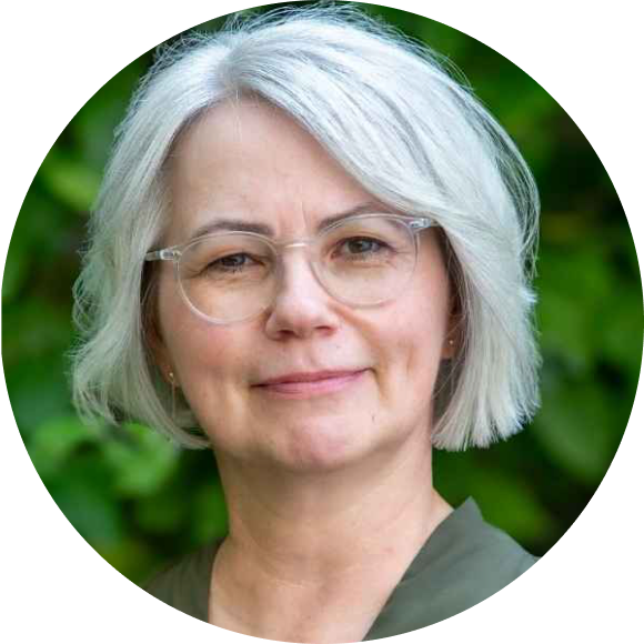 Eine Frau mit kurzen grauen Haaren und Brille lächelt sanft in die Kamera. Sie trägt ein grünes Oberteil und steht vor einem unscharfen Hintergrund aus grünem Laub.